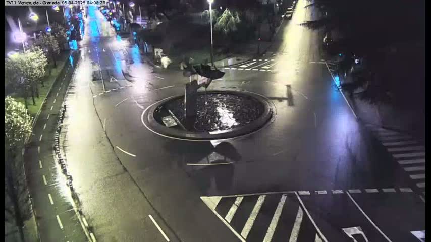 A wet roundabout with a central fountain is at the intersection of several roads at night.