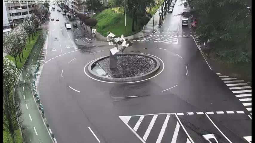 A wet roundabout with a modern sculpture in the center is surrounded by roads and trees on a rainy day.