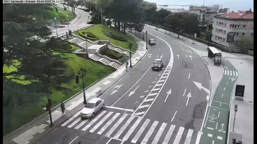 A curved road with several cars and a bus is seen from an aerial view, with a pedestrian crossing and a grassy area with stairs visible to the side, on an overcast day.