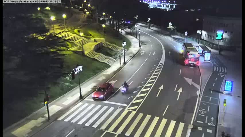 A nighttime street scene shows a red car and a motorcycle stopped at a crosswalk near a bus and stairs leading up a hill, all illuminated by streetlights.