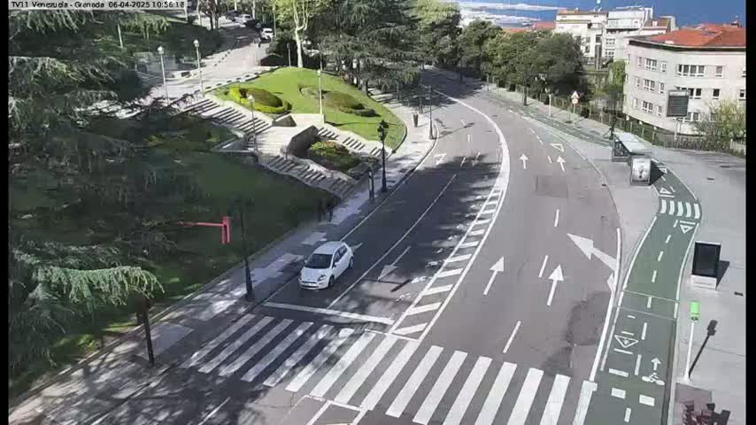 A white car is driving on a curved road that has a crosswalk and a bike lane next to it,  passing a park-like area with green landscaping under a sunny sky.