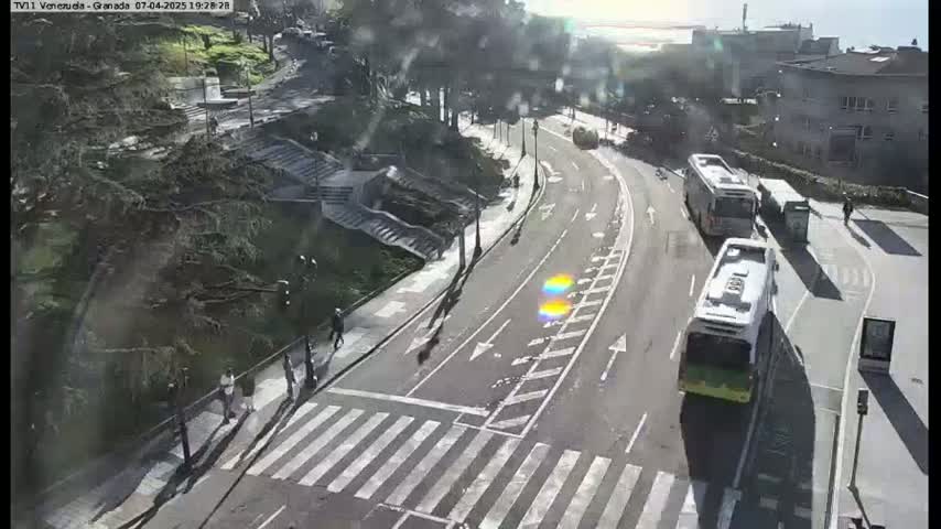 A sunny day reveals a road curving through a tree-lined area, with pedestrians crossing a crosswalk and several buses parked along the side.