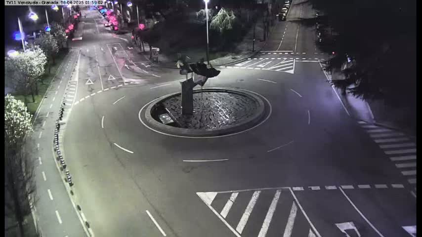 A nighttime aerial view shows a roundabout with a dark sculpture in the center, surrounded by roads with sparse traffic and pedestrian crosswalks.