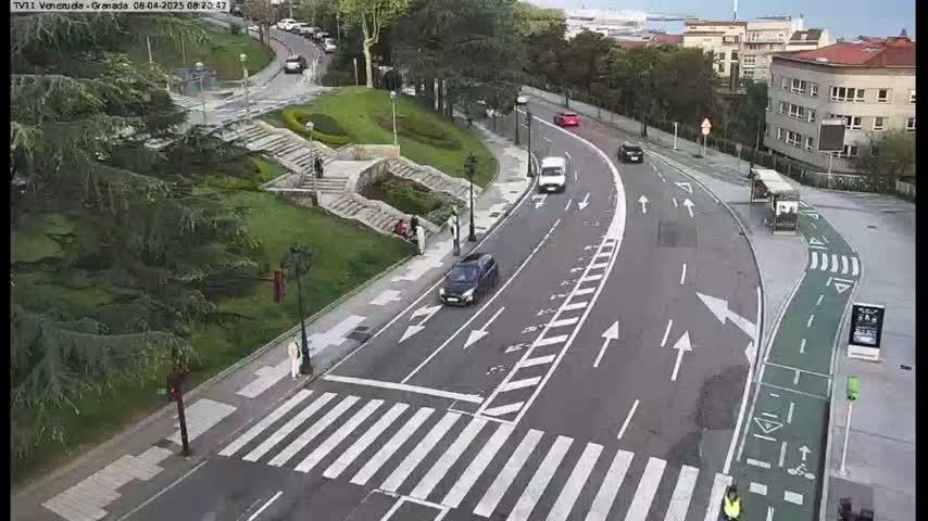 A paved road with a crosswalk curves through a landscaped area with stairs and greenery, under a mostly cloudy sky.