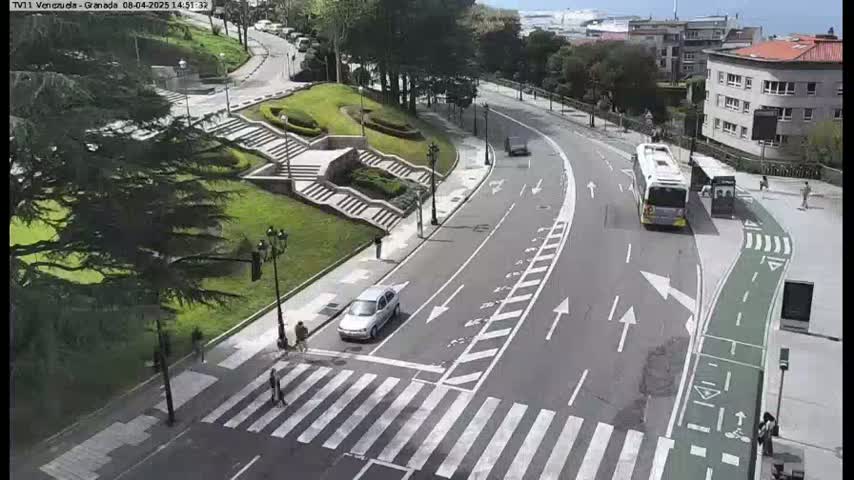 A sunny day reveals a curved road with a crosswalk, a bus stop, pedestrians, and a few vehicles, all situated near a grassy area with stone steps.