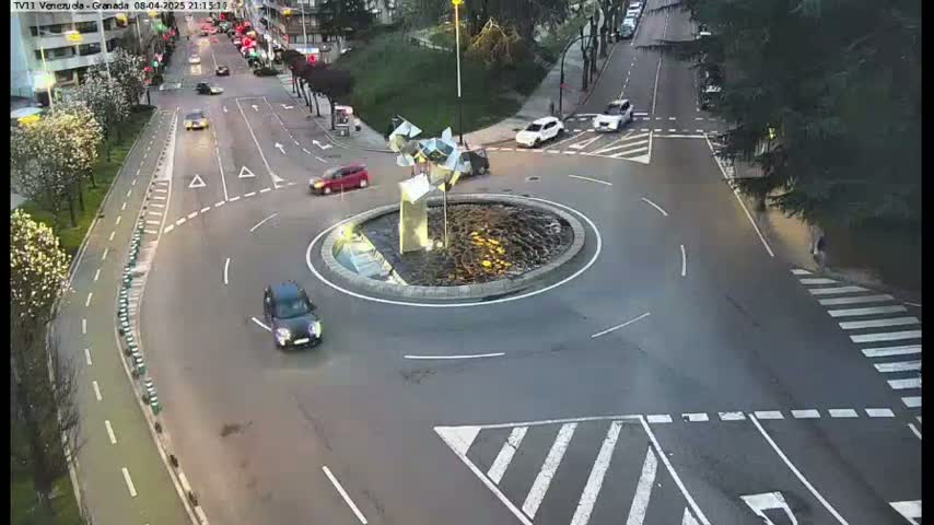 A high-angle, twilight view of a roundabout with several cars, a modern sculpture in the center, and surrounding roadways marked with crosswalks and bike lanes.