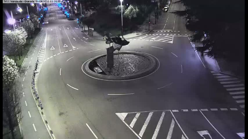 A nighttime aerial view shows a roundabout with a dark sculpture in the center, surrounded by roads and crosswalks.