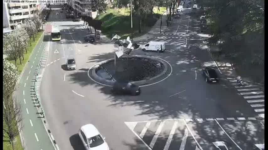 A street scene shows several cars driving around a roundabout with a metallic sculpture in the center, under a sunny sky.