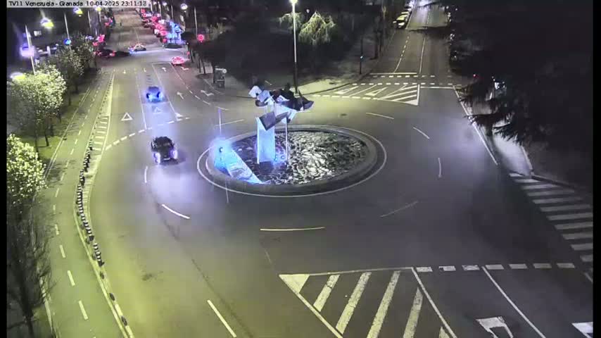 A nighttime aerial view of a roundabout with a few cars and a modern sculpture in the center.