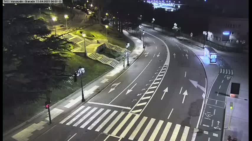 An empty, well-lit road curves through a nighttime cityscape, with pedestrian crosswalks and stairs leading to a park-like area.