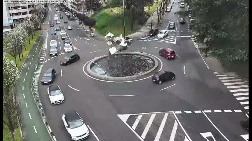 A roundabout with several cars circulating around a central fountain, on a partly cloudy day.