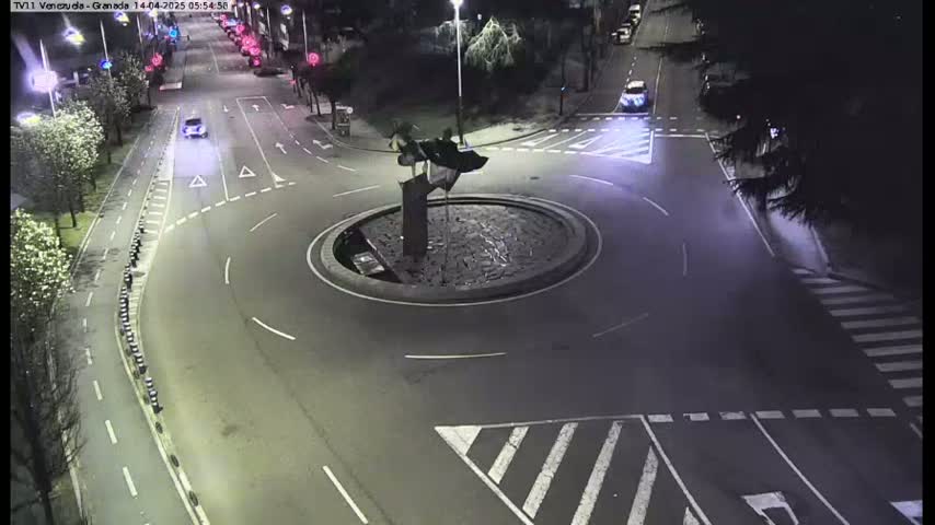 A nighttime aerial view shows a roundabout with a sculpture in the center, surrounded by roadways with cars and pedestrian crossings.