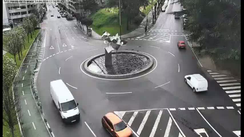 A wet roundabout with a modern sculpture in the center is surrounded by several cars and vans navigating the lanes on a partly cloudy day.