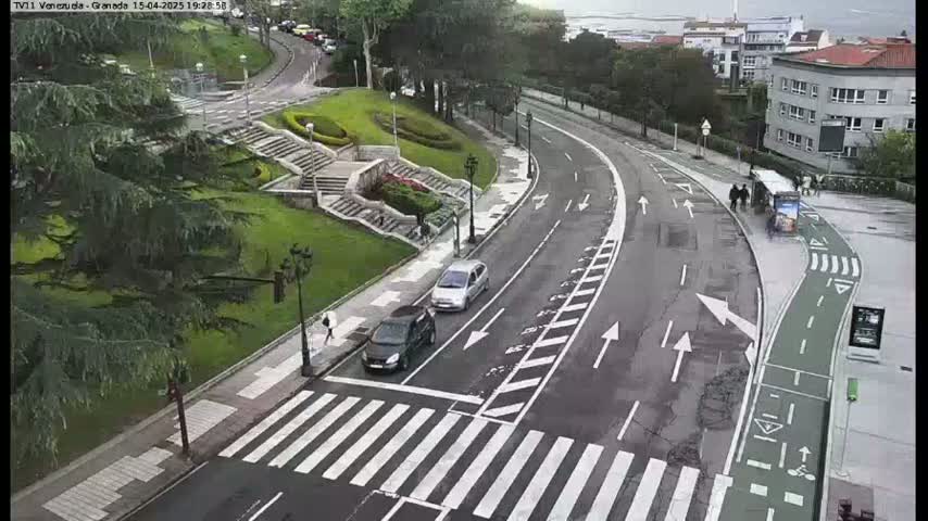 A wet, curved road with pedestrian crossings and bike lanes winds through a green space with stairs and buildings in the background.
