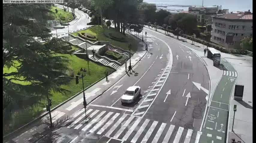 A curving road with a pedestrian crossing and bike lane winds through a landscaped area with stairs and trees, on a sunny day.