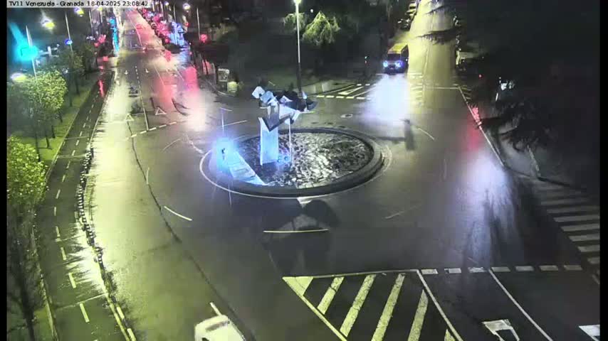 A wet, nighttime aerial view shows a roundabout with a modern sculpture in the center, surrounded by roads and vehicles.