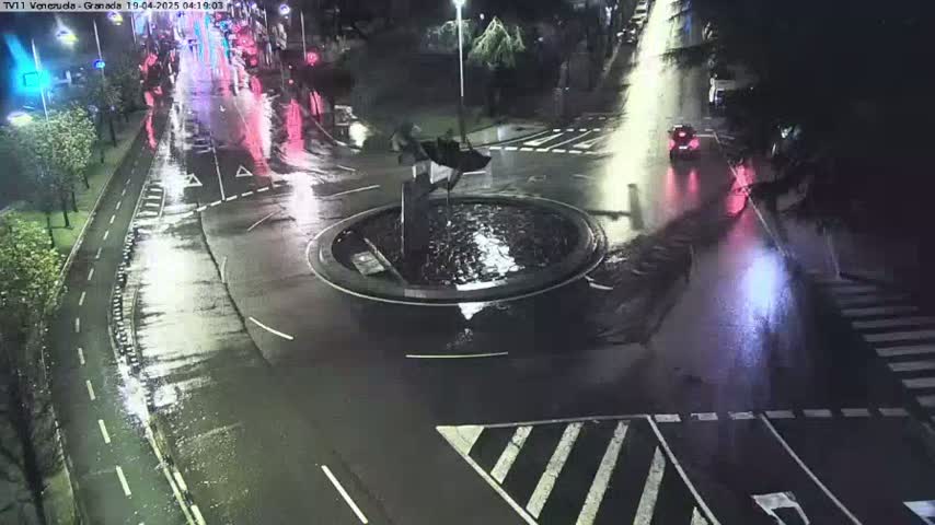 A wet roundabout at night with a few cars and a fountain in the center.