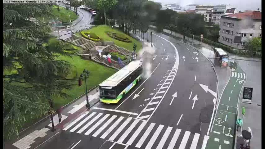 A green bus is driving on a wet, curved road next to a pedestrian crossing, passing by a bus stop and a grassy area with stairs.