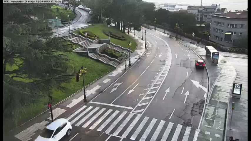 A wet road curves through a green space with stairs and buildings in the background under an overcast sky.