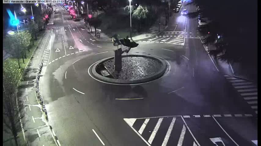 A nighttime aerial view shows a roundabout with a sculpture in the center, surrounded by wet roads and street markings.