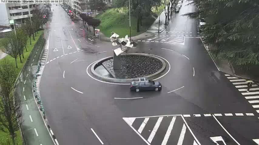 A wet, dark-colored car drives through a roundabout in the rain, near a modern sculpture in the center.
