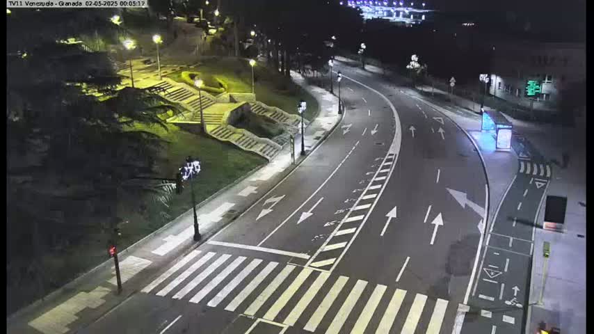 A nighttime, high-angle view shows a curving road with a crosswalk and streetlights illuminating a park-like area with stairs.