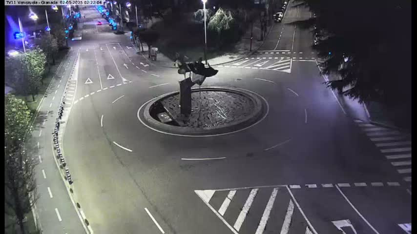 A nighttime overhead view shows a mostly empty roundabout with a dark sculpture in the center, surrounded by roads with light markings and pedestrian crossings.