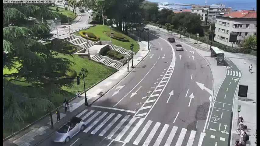 A sunny day shows a curving road with a crosswalk, pedestrian traffic, and several cars, next to a grassy area with stairs and landscaping.