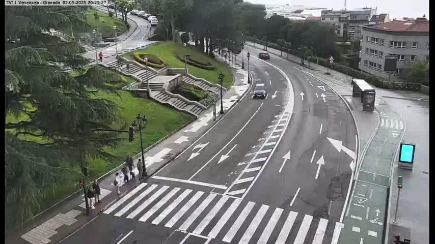 A curved road with pedestrian crossings and a few people is seen from above on an overcast day, next to a grassy area with stairs and trees.