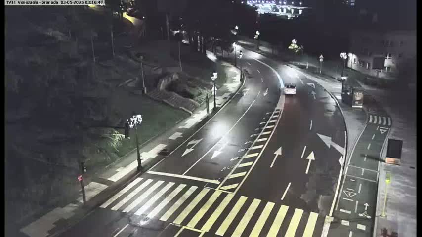 A wet, nighttime street scene shows a curving road with a crosswalk, streetlights, and a single car.