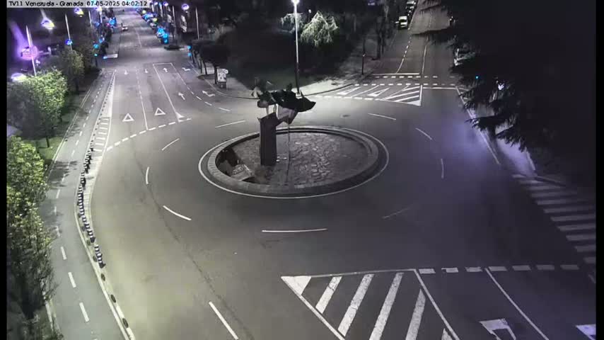A nighttime aerial view shows a roundabout with a dark sculpture in the center, surrounded by streets with crosswalks and minimal traffic.