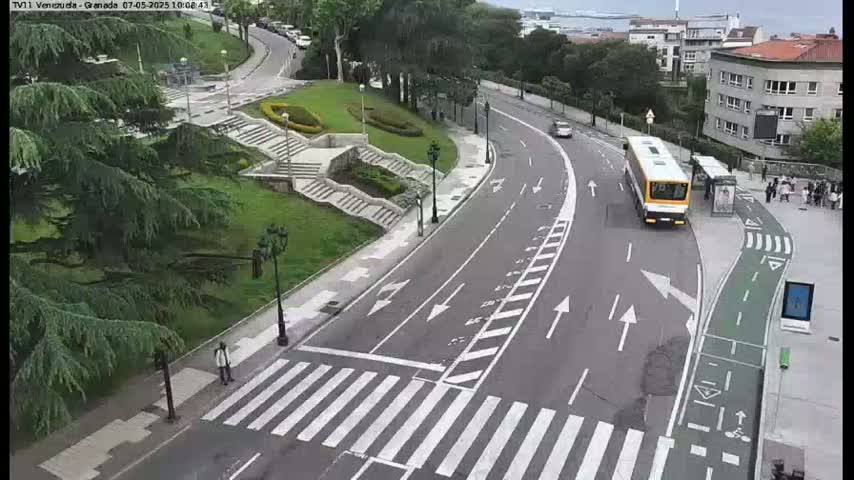A paved road curves through a landscaped area with a bus and pedestrians, under overcast skies.