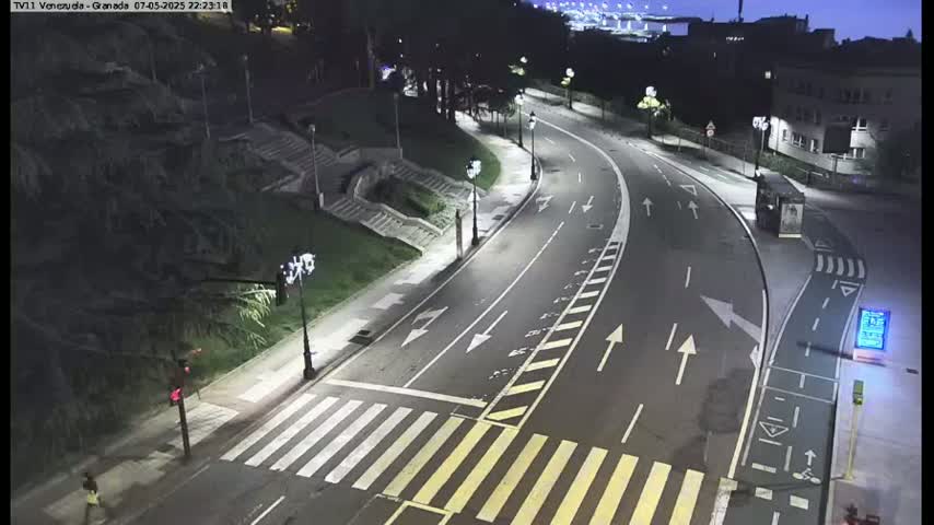 A mostly empty, curved road at night is illuminated by streetlights, with a pedestrian crossing and a person walking in the distance.