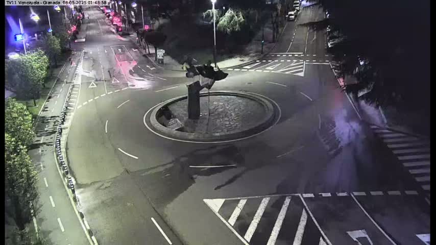 A nighttime overhead view shows a roundabout with a dark sculpture in the center, surrounded by dark asphalt roads with crosswalks.