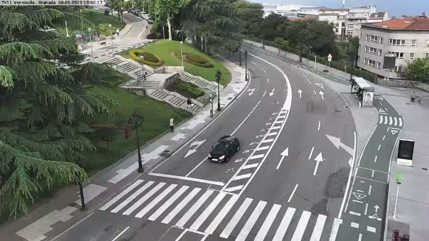 A curving road with a crosswalk and a dark-colored car is seen from an elevated viewpoint, on an overcast day, with greenery and buildings in the background.
