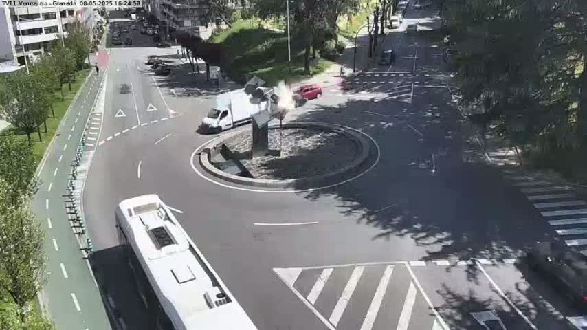 A sunny day shows a roundabout with a sculpture in the center, several vehicles including a bus and van, and a bike path alongside a road.
