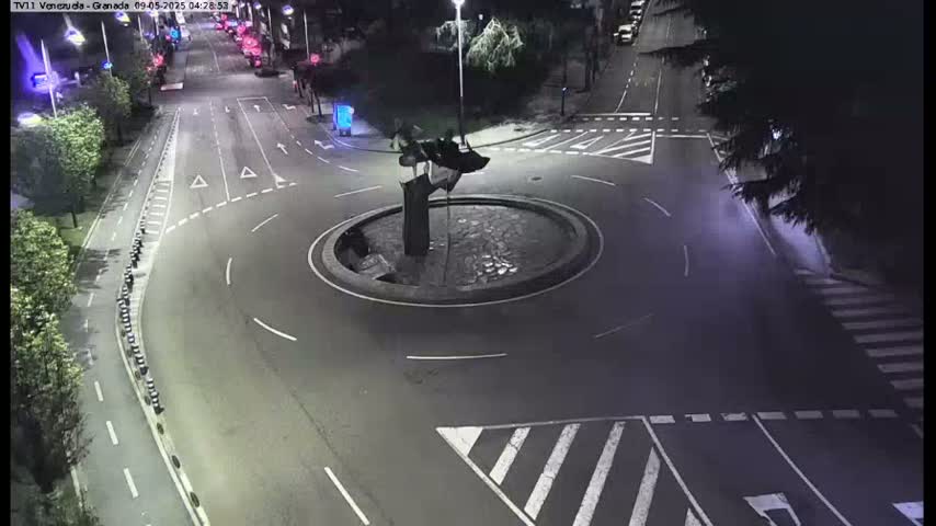 A nighttime aerial view of a roundabout with a dark sculpture at its center, surrounded by roads with minimal traffic.