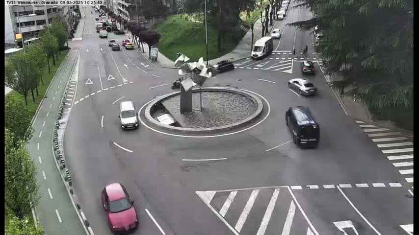 Several cars navigate a roundabout with a modern sculpture in the center, under a mostly clear sky.
