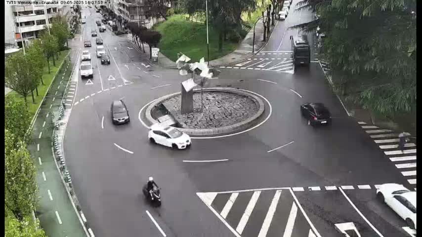 A wet roundabout with several cars and a motorcycle, surrounded by roads and green spaces.