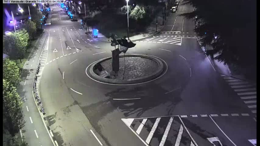 A nighttime aerial view shows a roundabout with a dark sculpture in the center, surrounded by dark roads and street markings.