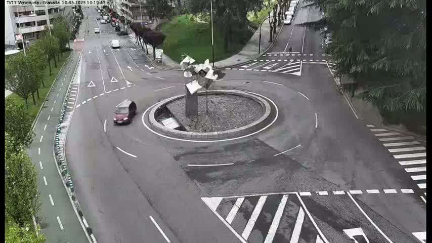 A wet roundabout with a modern sculpture in the center is surrounded by a roadway with cars and a bike path.