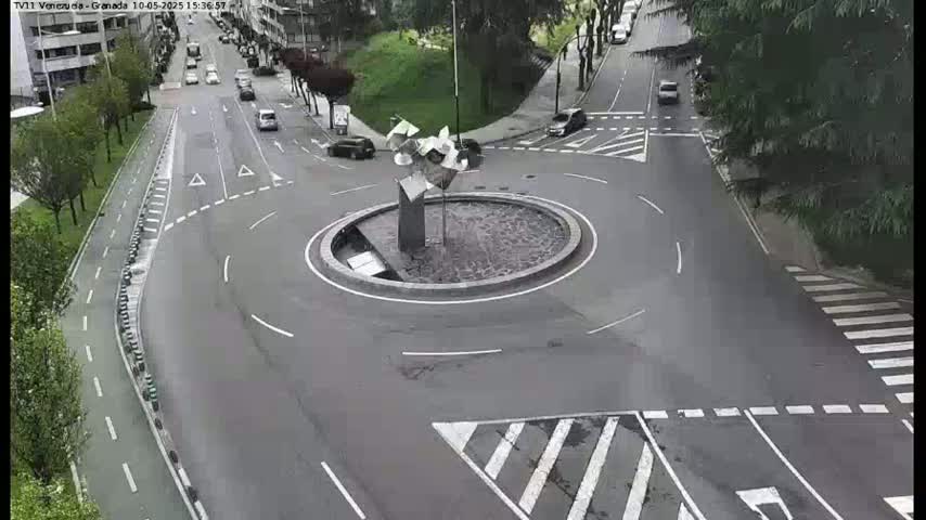 A roundabout with a modern sculpture in the center is surrounded by roads with a few cars and lush green trees, under overcast skies.