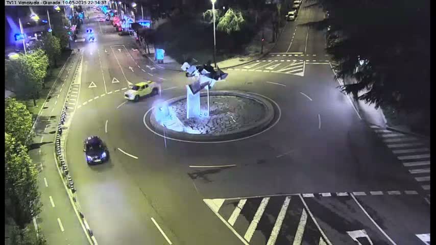 A nighttime aerial view shows several cars navigating a roundabout with a modern sculpture in the center.