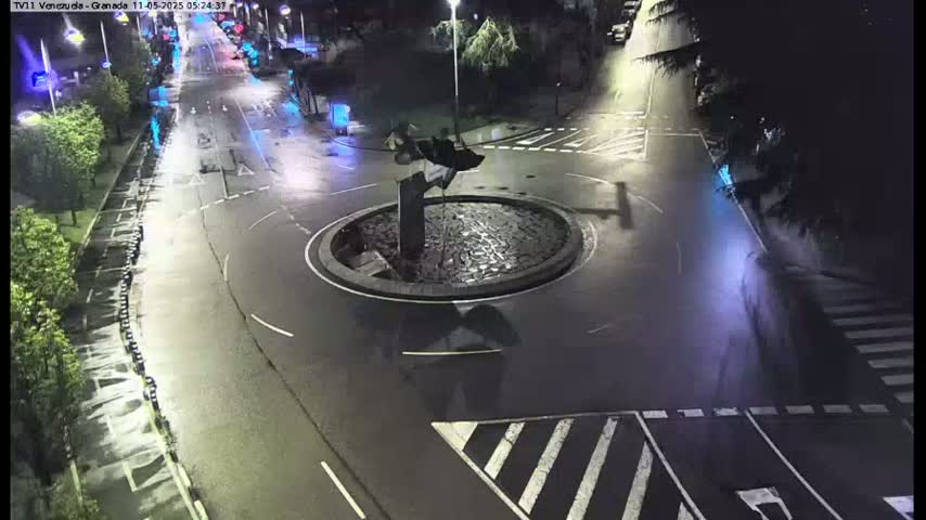 A nighttime aerial view shows a roundabout with a dark sculpture in the center, surrounded by wet roads and sparse traffic.