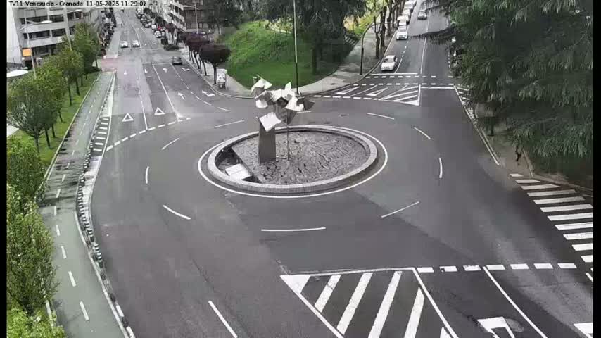 A wet roundabout with a modern metal sculpture in the center is surrounded by roads with cars and pedestrian crossings.
