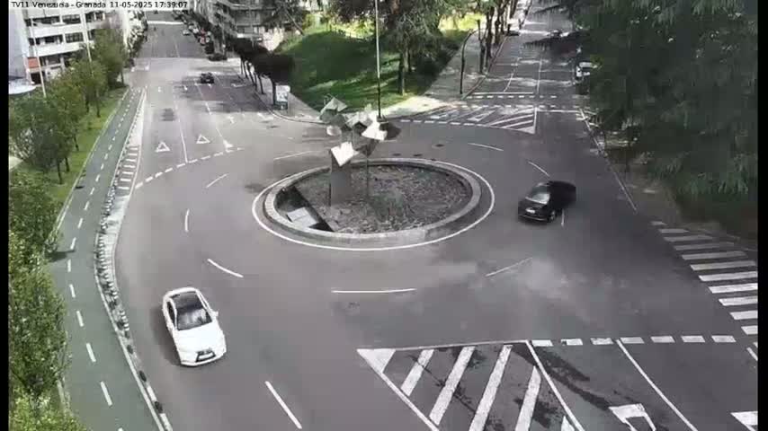 A roundabout with a sculpture in the center is surrounded by roadways and a pedestrian crossing, with two cars visible on the roads under a partly sunny sky.