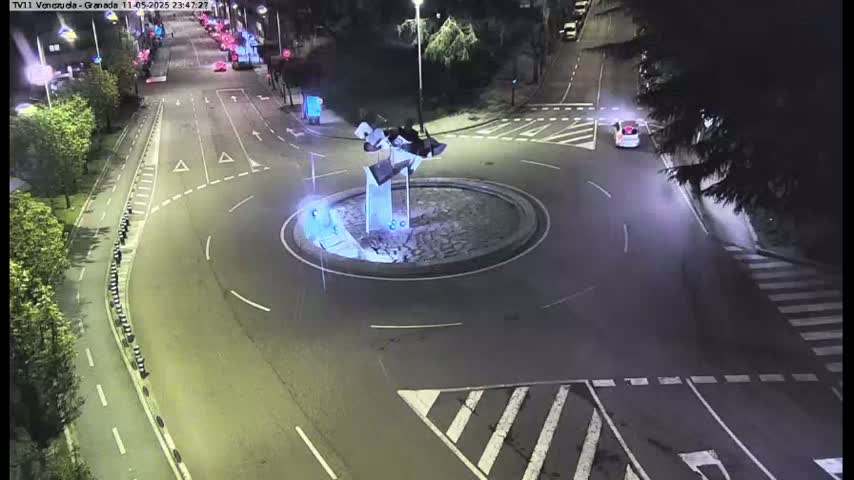 A nighttime aerial view shows a roundabout with a modern sculpture in the center, surrounded by roads with minimal traffic under artificial light.
