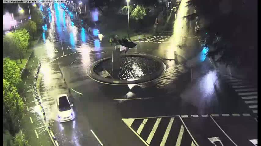 A nighttime, rain-soaked roundabout with a central fountain, a white car driving through it, and surrounding roadways showing wet pavement and street markings.