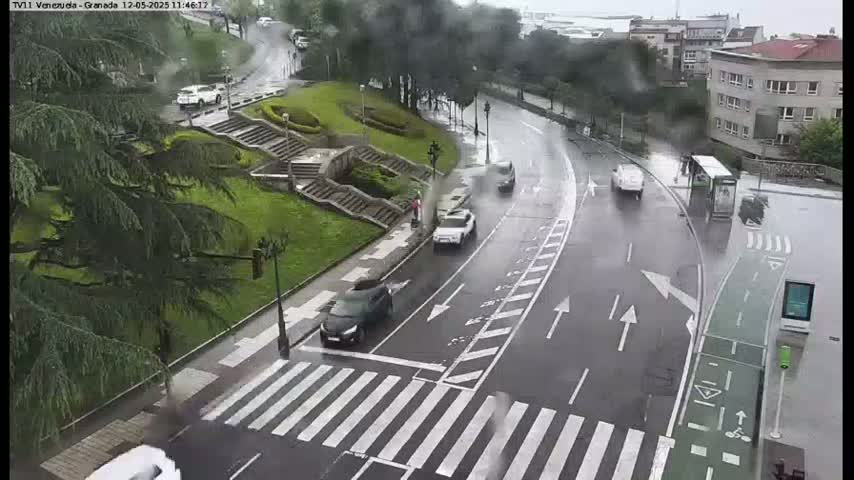 A rain-slicked street curves past a pedestrian crossing and a set of stairs leading to a grassy area, with several cars and a bus visible.