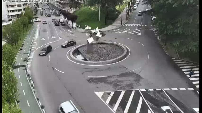 An aerial view of a roundabout with several cars driving around it, on a mostly sunny day.
